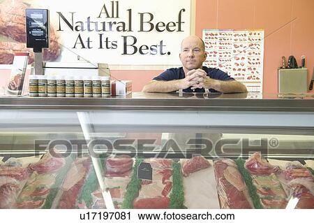 Stock Image - Supermarket employee stands at meat counter in supermarket. Fotosearch