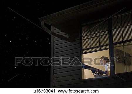 Stock Image - Teenage boy stands with telescope at open window looking at night sky. Fotosearch