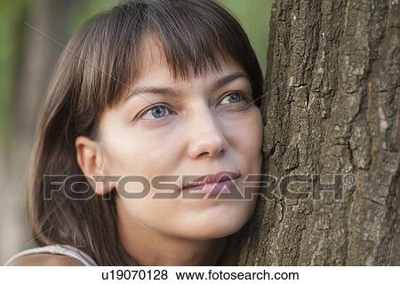 Stock Photo - Woman leans on tree trunk with a faraway look in her eyes. Fotosearch