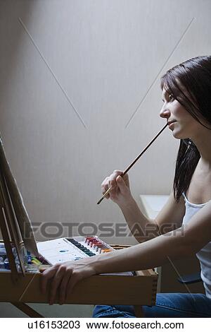 Stock Image - Woman sitting in front of easel and holding paintbrush. Fotosearch