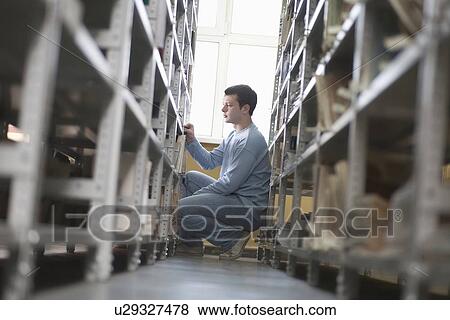 Stock Photo - Young man crouches at library shelf. Fotosearch