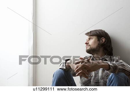 Stock Image - Young man in checked shirt sits with hands clasped against a wall. Fotosearch