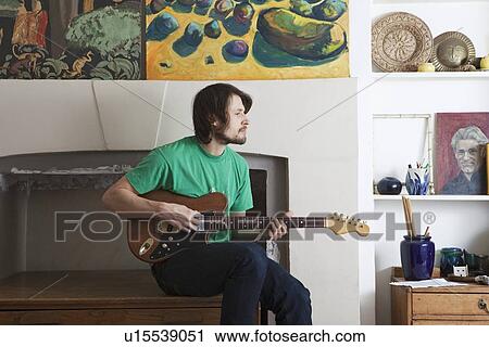Stock Image - Young man sits in living room playing electric guitar. Fotosearch