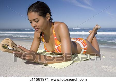 Picture - Young woman in retro swimwear lies on beach reading a book. Fotosearch