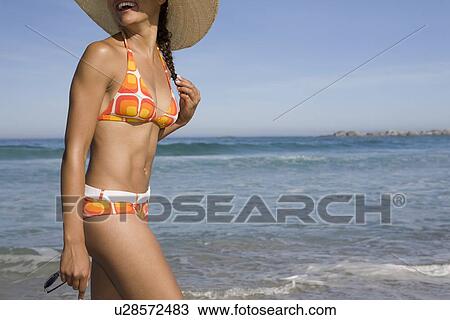 Stock Image - Young woman in sunhat and retro swimwear walking on beach. Fotosearch