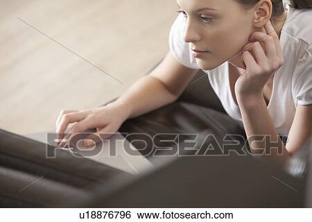 Stock Photograph - Young woman lies on leather sofa working on laptop. Fotosearch