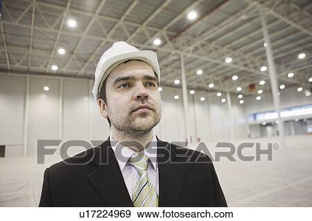 Stock Photo - Businessman in hard hat stands in brightly lit warehouse. Fotosearch