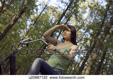Stock Image - Young woman stands shielding eys with bicycle in woodland. Fotosearch