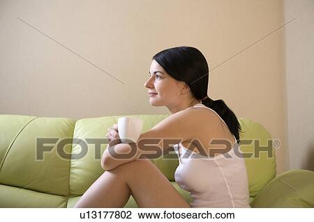 Young woman sits on sofa, side view holding cup View Large Photo Image Stock Image - Young woman sits on sofa, side view holding cup. Fotosearch