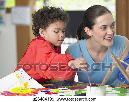 Stock Photography - Art teacher squatting next to boy painting in art class. Fotosearch