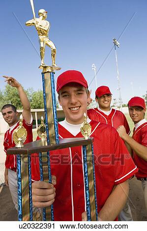 Stock Image - Baseball player holding trophy on field, team-mates in background. Fotosearch