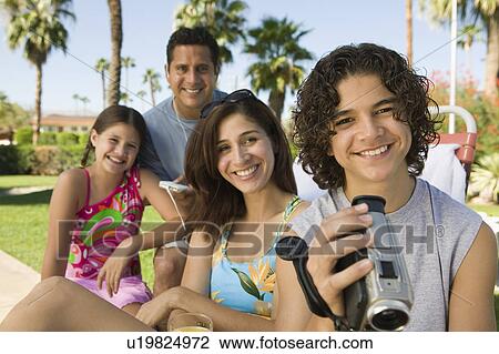 Stock Image - Boy (13-15) holding camcorder, sitting outdoors with sister (7-9) and parents, portrait.. Fotosearch