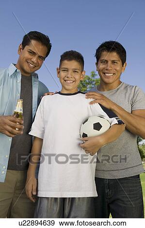 Stock Photo - Boy (13-15) holding soccer ball, standing outdoors with two brothers, front view.. Fotosearch
