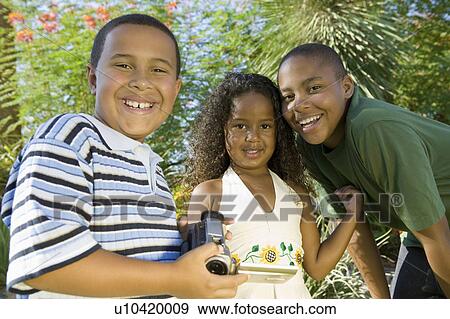 Stock Photo - Boy (7-9) holding camcorder, with younger sister (5-6) and older brother (10-12), portrait.. Fotosearch