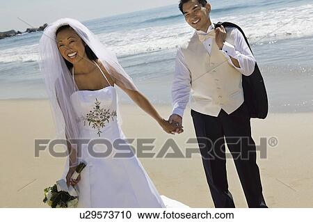 Stock Image - Bride and Groom holding hands on beach. Fotosearch