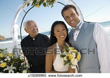 Bride and Groom with priest under archway at ocean, (portrait) View Large Photo Image Stock Photography - Bride and Groom with priest under archway at ocean, (portrait). Fotosearch