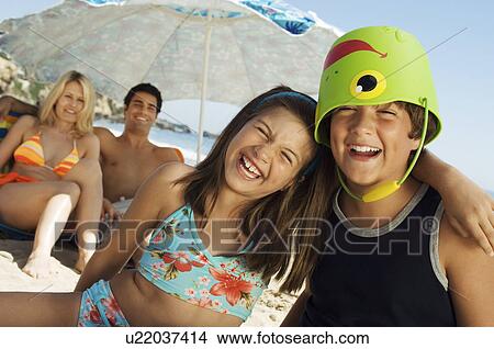 Picture - Brother and sister sitting on beach, arm around, boy with bucket on head. Fotosearch
