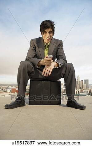 Stock Photo - Business man sitting on briefcase using mobile phone in city. Fotosearch