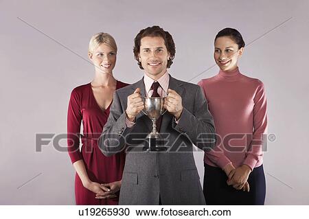 Stock Image - Businessman holding trophy with female colleagues, portrait. Fotosearch