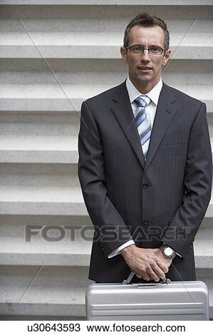 Stock Image - Businessman standing, holding briefcase in front of wall, portrait. Fotosearch