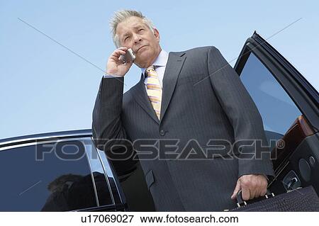 Businessman standing in open door of car, talking on mobile, low angle view View Large Photo Image Stock Photo - Businessman standing in open door of car, talking on mobile, low angle view. Fotosearch
