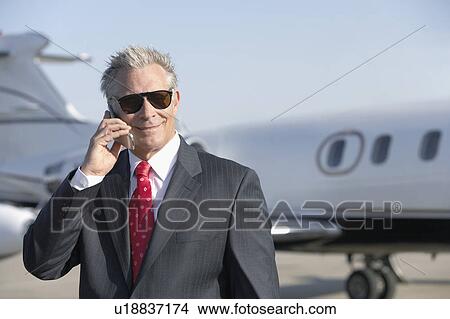 Picture - Businessman standing on landing strip near private jet, talking on mobile. Fotosearch