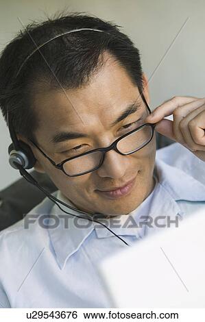 Stock Photograph - Businessman wearing headset, using laptop, (close-up). Fotosearch