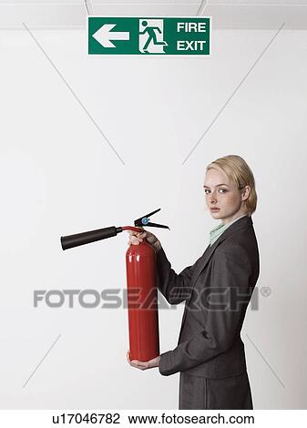 Stock Image - Businesswoman holding fire extinguisher under exit sign. Fotosearch
