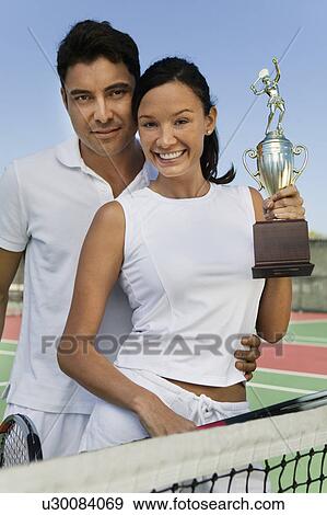 Stock Photo - Couple by net at tennis court holding trophy, portrait. Fotosearch