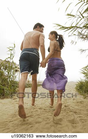 Stock Image - Couple Holding Hands, Walking on Beach, back view. Fotosearch