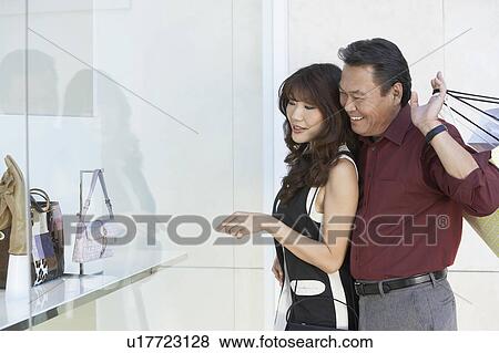 Stock Photo - Couple looking at merchandise in shop window, arm around, holding shopping bags. Fotosearch