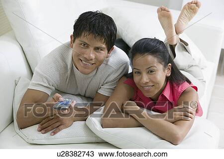 Stock Image - Couple lying on Couch with Remote Control, front view. Fotosearch