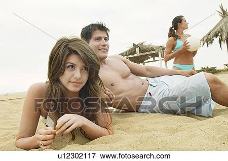 Stock Photo - Couple Lying on Sand by Friend with Volleyball. Fotosearch
