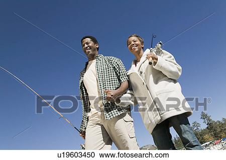 Stock Image - Couple on fishing trip, holding hand, smiling, (low angle view). Fotosearch