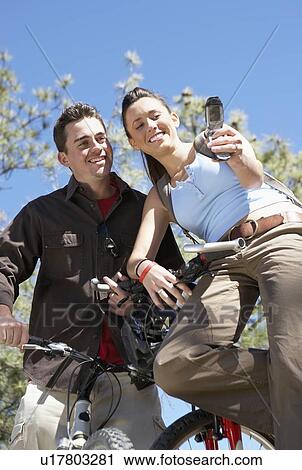 Stock Image - Couple on Mountain Bikes using mobile phone, photographing selves, low angle view.. Fotosearch