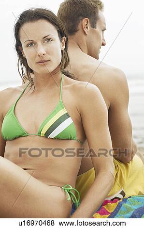 Stock Photo - Couple sitting back to back on beach, portrait of woman. Fotosearch