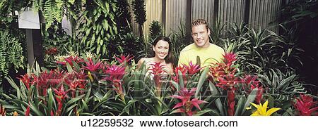 Stock Image - Couple standing among plants in plant nursery, portrait. Fotosearch
