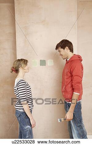 Stock Photo - Couple standing face to face, choosing paint colour samples on wall. Fotosearch
