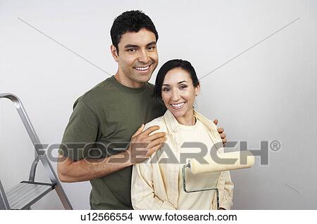 Picture - Couple standing in unpainted room with paint roller, portrait. Fotosearch