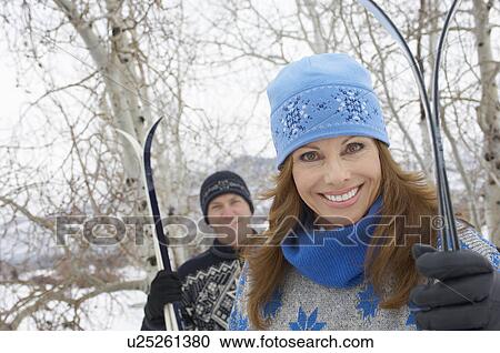 Stock Image - Couple standing outdoors, holding skis in snow. Fotosearch