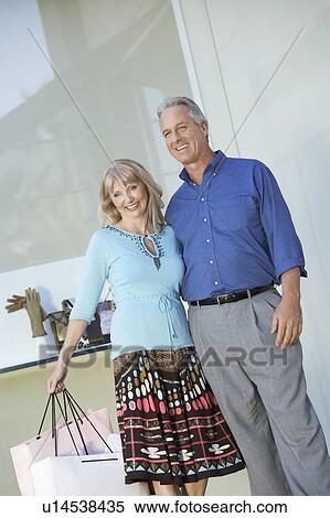 Couple standing outside shop, arm around each other, holding shopping bag View Large Photo Image Stock Photography - Couple standing outside shop, arm around each other, holding shopping bag. Fotosearch