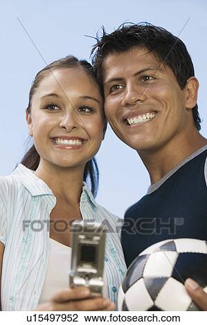 Stock Image - Couple using mobile phone, man holding soccer ball.. Fotosearch