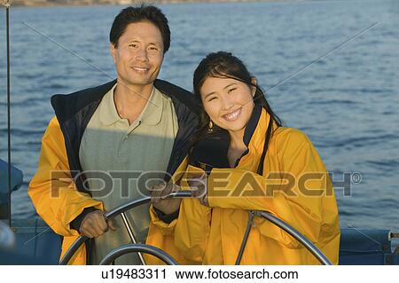 Stock Image - Couple wearing yellow anoraks at steering wheel of sailboat, (portrait). Fotosearch