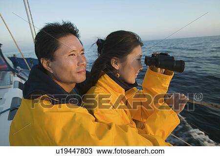 Stock Image - Couple wearing yellow anoraks on yacht, woman looking through binoculars. Fotosearch