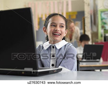 Stock Image - Elementary schoolgirl sitting by laptop in classroom, smiling. Fotosearch