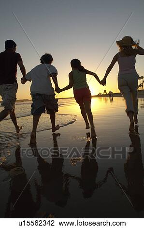 Stock Image - Family running through sea at sunset, back view, silhouette. Fotosearch
