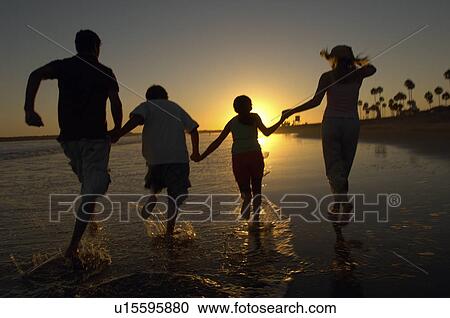 Stock Image - Family running through sea holding hands at sunset, back view, silhouette. Fotosearch