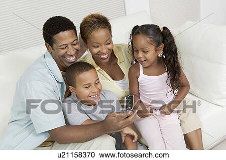 Stock Image - Family sitting on sofa looking at picture on camera phone. Fotosearch