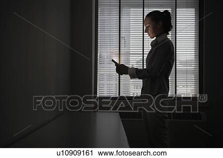 Stock Image - Female office worker standing using mobile phone in darkened office, side view. Fotosearch