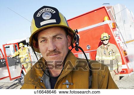 Stock Photograph - Fire fighter standing in front of colleagues and fire engine. Fotosearch